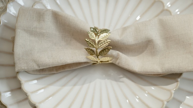 A golden leaf napkin ring encircles a linen napkin sitting on a white porcelain plate.