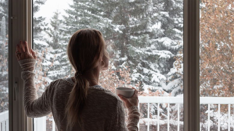 woman holding mug looking out window at snow outside