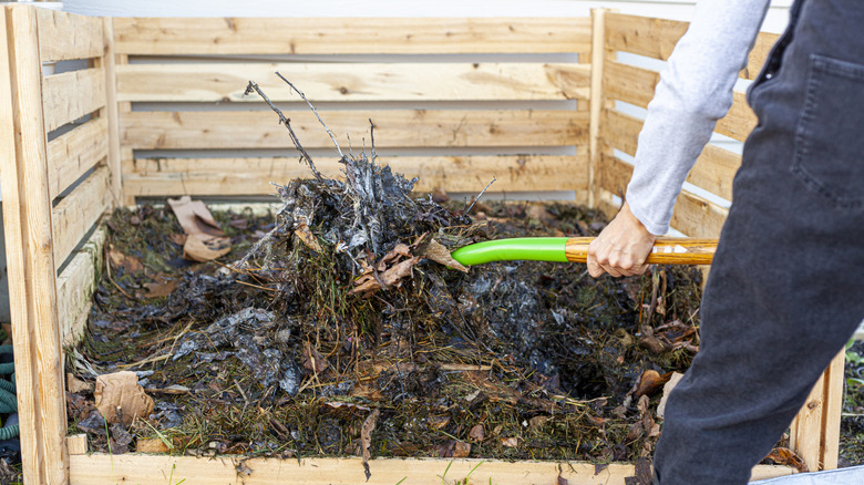 A gardener turns compost in a large wooden bin with a shovel.