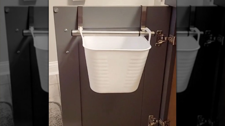 A small plastic basket with handles hangs on a towel rod on the back of a bathroom cabinet door
