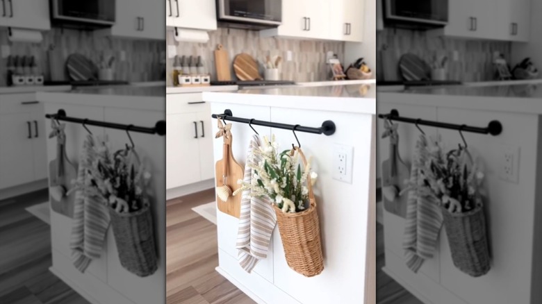 A cutting board, towel, and decorative basket hang from a towel bar on the side of a kitchen island