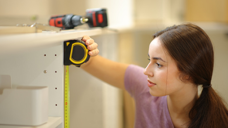 Woman holding a tape measure case up against the corner of a cabinet with the tape extended towards the floor