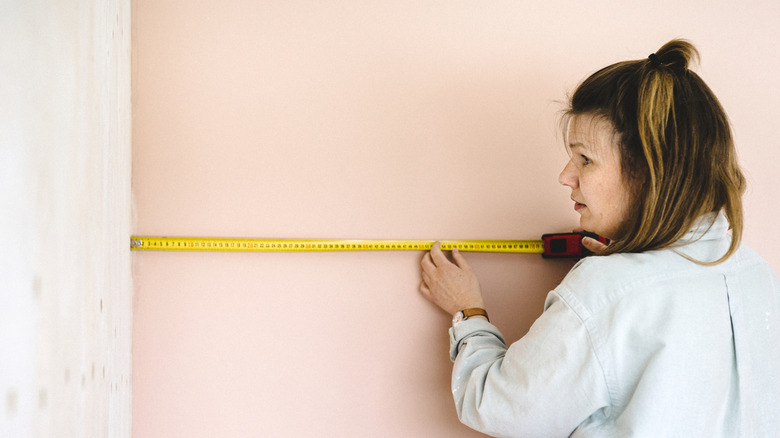 Woman measuring the corner of a room with one light pink wall and one wall painted with primer