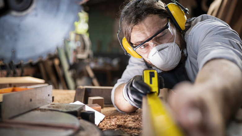 Worker wearing sound protecting ear muffs and respirator mask in a wood shop measuring a wooden beam with a yellow and black measuring tape