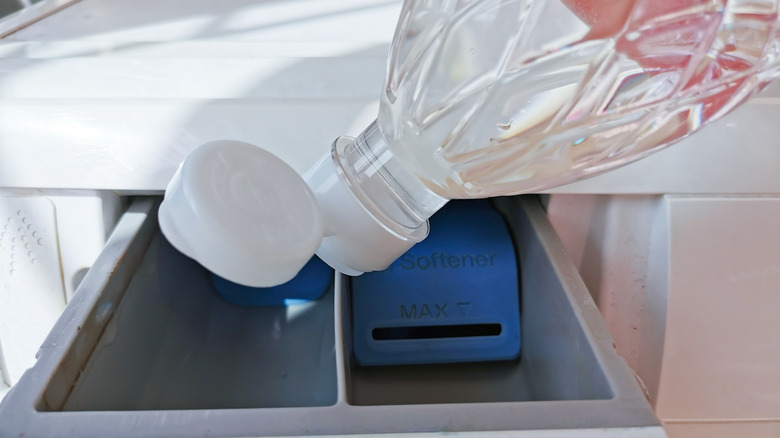 A person pours vinegar from a clear bottle into the drawer of a laundry machine