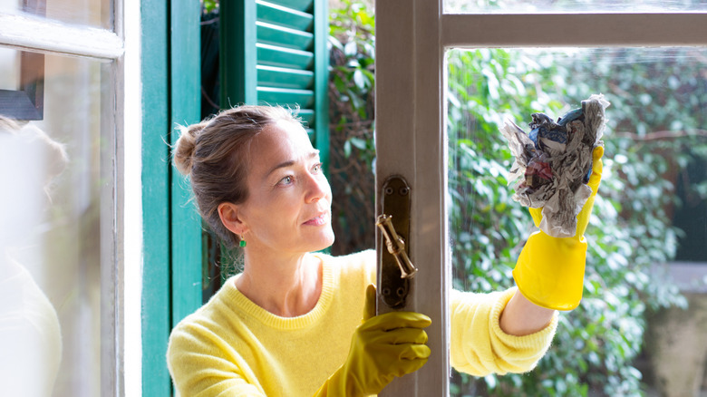 A woman in a yellow shirt cleans glass windows with newspaper while smiling