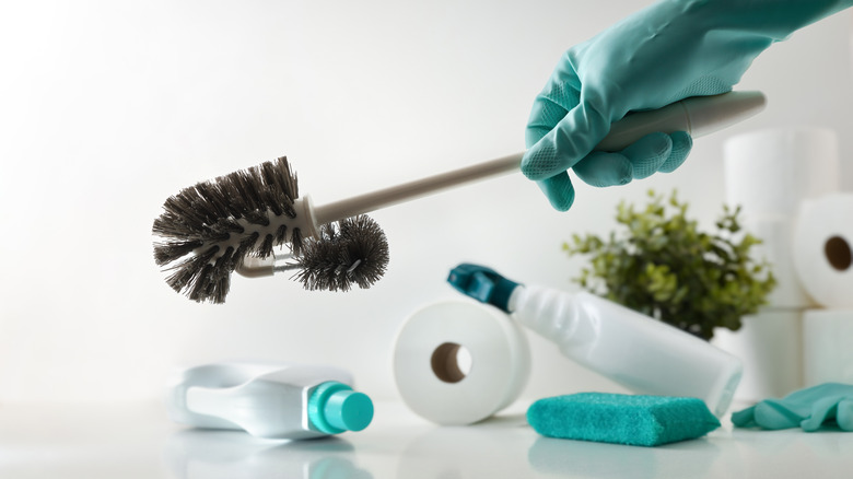A gloved hand holds a cleaning brush with many other cleaning products in the background, including cleaning sprays and sponges