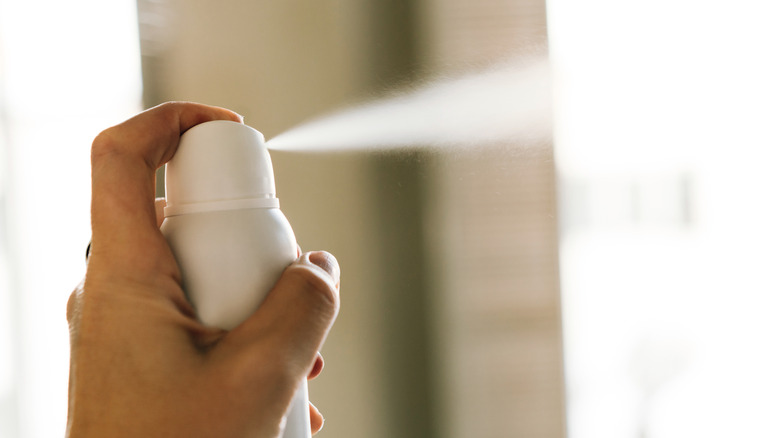 A hand presses down on the top of a white hairspray bottle, spraying white mist