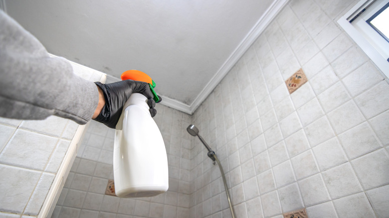 A hand holds a white bottle, ready to spray it onto bathroom shower tile