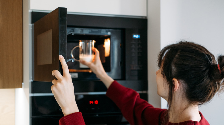 A woman puts a jug of milk into a microwave that is mounted into the wall.