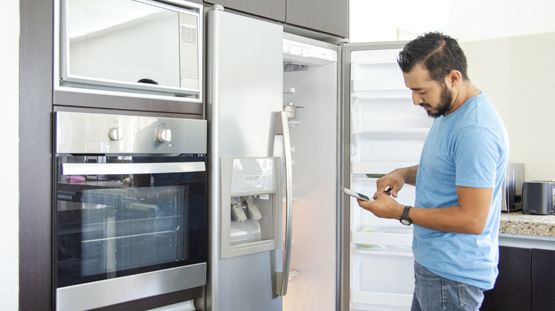 Man stands in front of the open door of a side-by-side door fridge while looking at his smartphone.