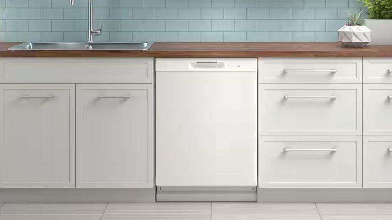 A compact white dishwasher in a kitchen with white cabinets and drawers