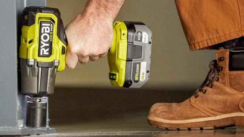 A person wearing workboots uses Ryobi's High Torque Impact Wrench on a metal beam on the ground.