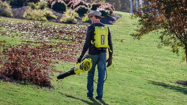 A man wears the backpack battery and holds the leaf blower of this Ryobi tool as he blows leaves off of a green lawn.