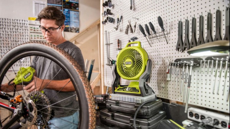 A man works on a bike in the garage with the Ryobi portable fan behind him.