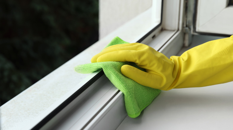 A hand in a yellow glove cleans a metal window frame with a green cloth.