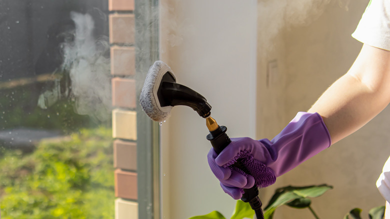 A person wearing purple gloves holds a steam cleaner to a window.
