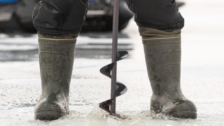 A man in boots drilling an ice auger into ice.