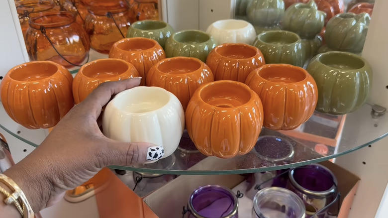 A person with a cow print painted nail and gold bracelets holding a white pumpkin candle holder in front of orange and green ones