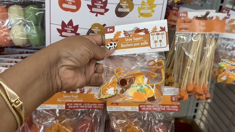 A person with a cow print-painted thumbnail and gold bracelets holding a pack of fall-themed straw toppers at Dollar Tree