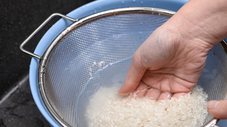 A person rinses rice in a plastic bowl full of water using a metal sieve.