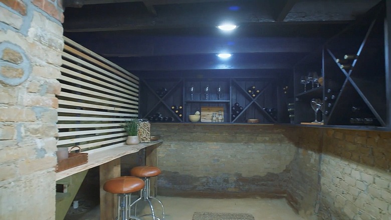 A wine cellar with exposed brick walls and painted black wooden shelves holding bottles of wine and glasses.