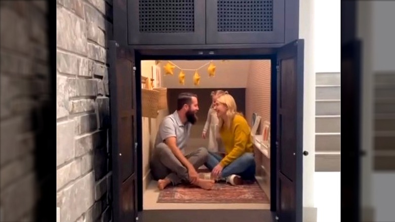 Celebrity designer Jasmine Roth, her husband and daughter hang out inside a secret playroom hidden behind a dresser with small table, chairs, shelf, and rug.