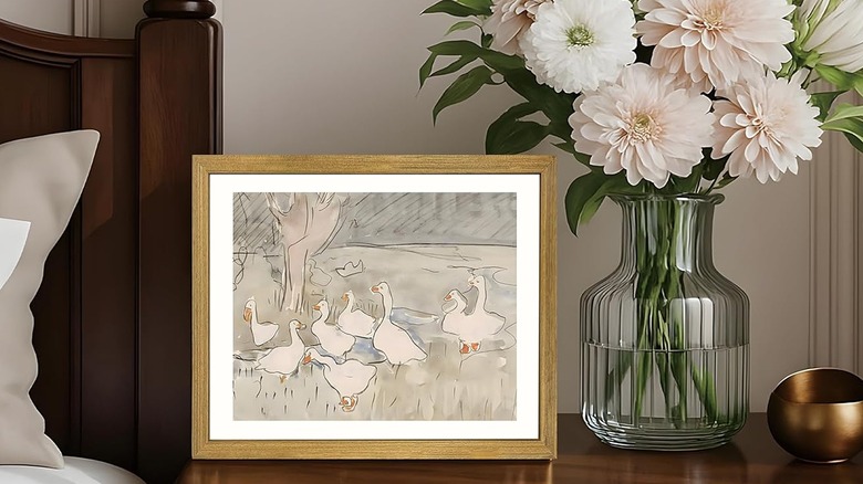 A framed watercolor painting of geese in the countryside sits on a nightstand next to a vase of flowers.