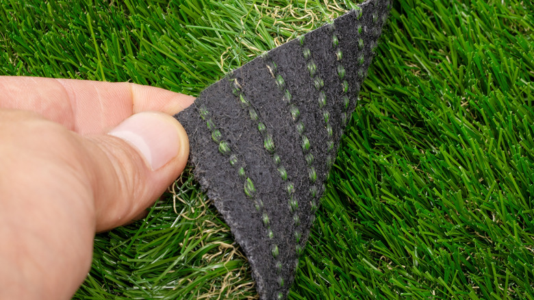 A hand peeling back artificial turf to examine synthetic backing