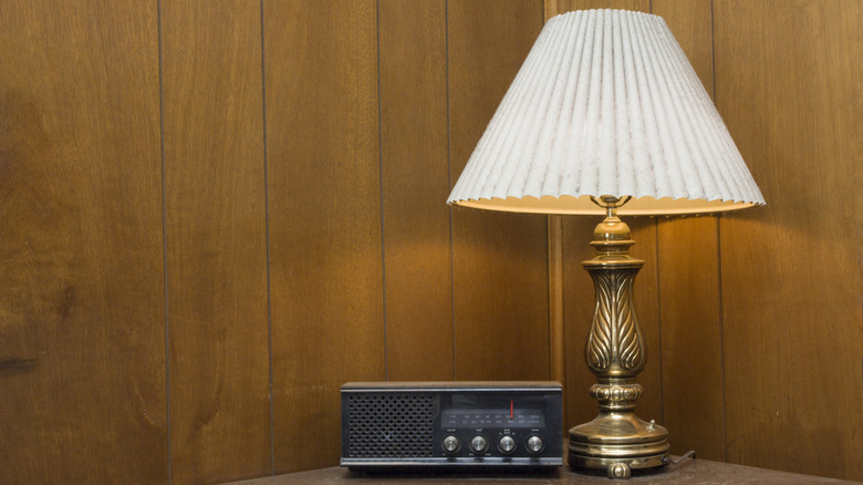 A brass table lamp on a table with a vintage clock radio in a room with wood paneling.