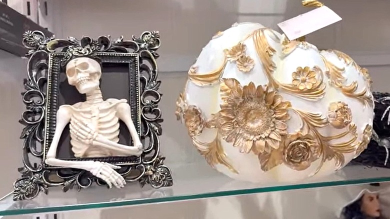 A skeleton in a frame, a pumpkin decorated with gold flowers, and a black wreath with spiders on the shelf in a store.