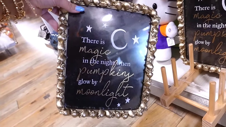 A hand holds a gold frame with skulls. Inside there's a poster with a message about pumpkins.