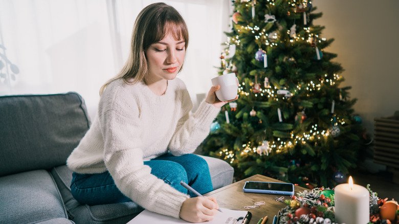 Woman with coffee in hand making a list on a clipboard with a Christmas tree in the background
