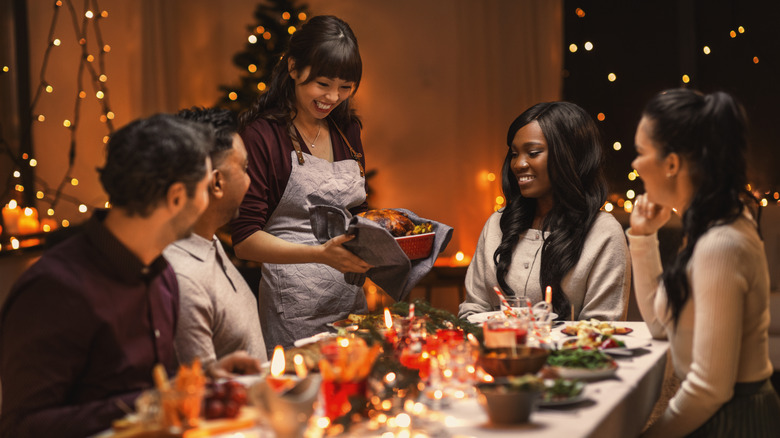 Dinner party with Christmas lights, woman holding a pan with roasted turkey, two men and two women sitting at a festive table