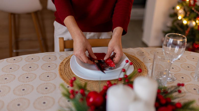 Woman placing napkins on place setting for a Christmas dinner, with lit Christmas tree in background