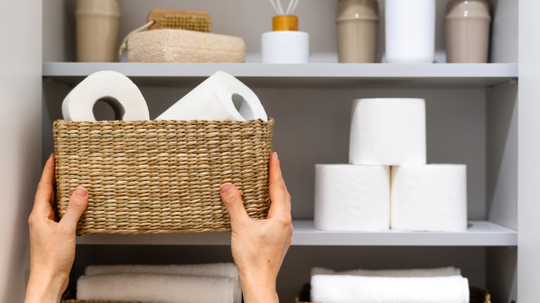 Person holding basket of toilet paper rolls near shelf of bathroom supplies