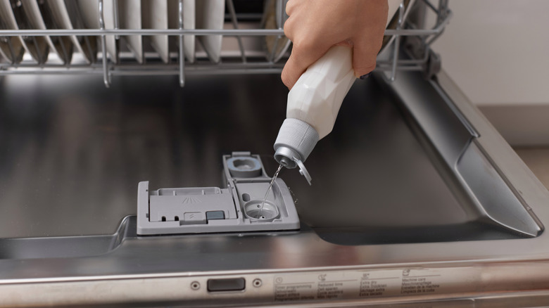 Hand holding bottle of rinse aid, pouring it into dish washer.