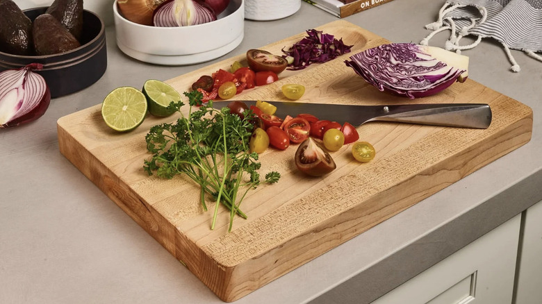A wood cutting board being used to prep tomatoes, cilantro, limes, and cabbage