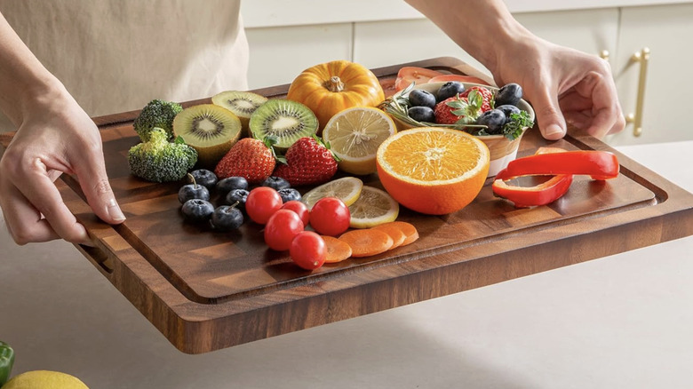A person presenting a variety of fruits and vegetables on a walnut cutting board