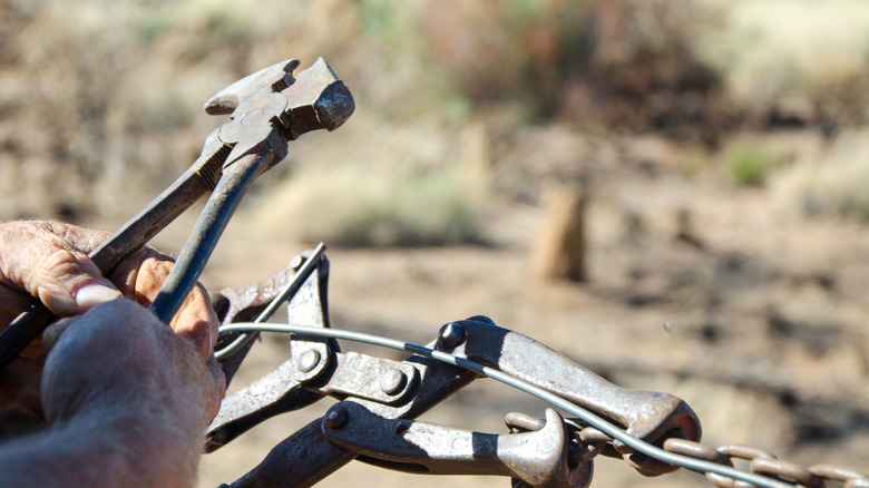 A set of hands handling an old fence plier tool above a piece of wire outside.