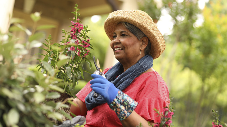 A smiling older woman with dark skin prunes her flower garden.