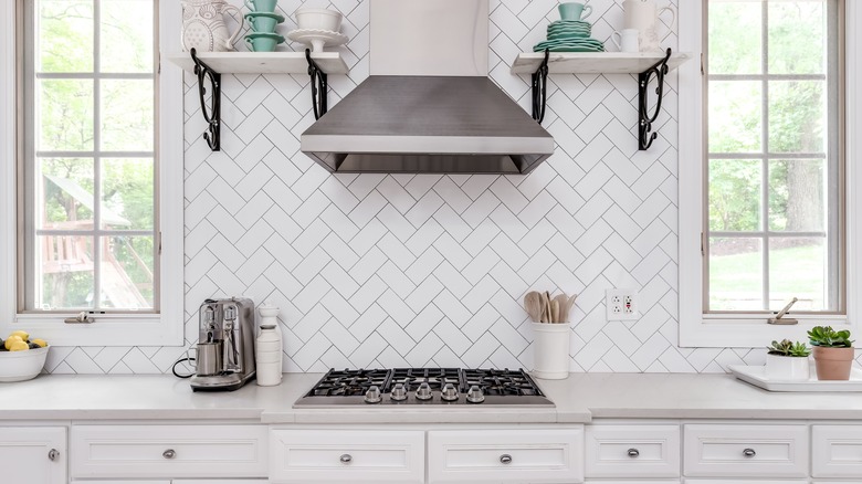 A kitchen with shelves on white herringbone tile backsplash, white cabinets, and stainless steel appliances.
