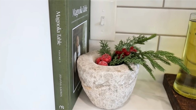 A stone mortar on top of kitchen countertop is filled with pine tree cuttings and red berries.