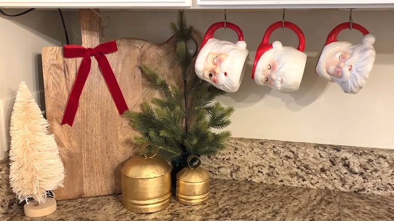 Three Santa mugs hang from hooks underneath cabinet in kitchen with granite countertops.