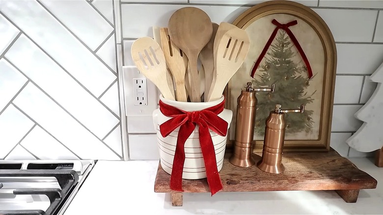 Ceramic utensil holder decorated with a bow made from ribbon on top of a wooden platform next to salt and pepper mills and a Christmas tree painting.