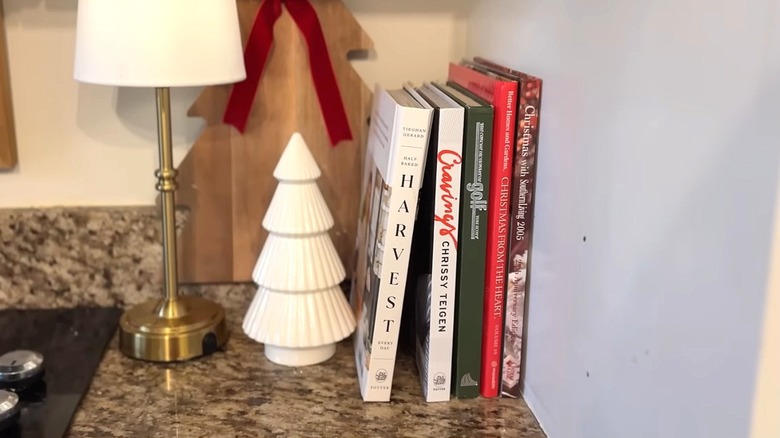 A collection of cook books rests against the wall in front of tree-shaped board and white ceramic tree figurine.