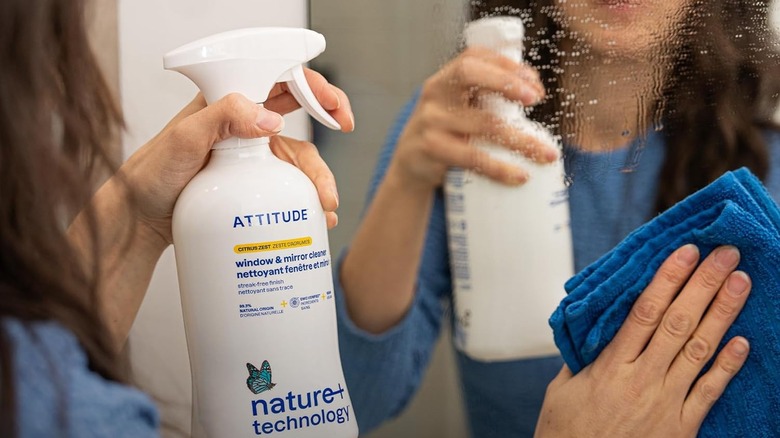 A woman uses a blue cloth to wipe a mirror sprayed with Attitude Window & Mirror Cleaner
