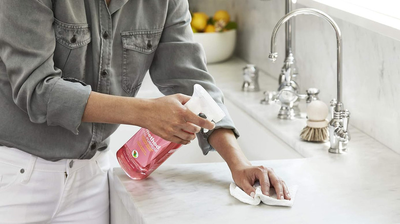 A woman sprays a bottle of Method All-Purpose Cleaner on a white kitchen countertop