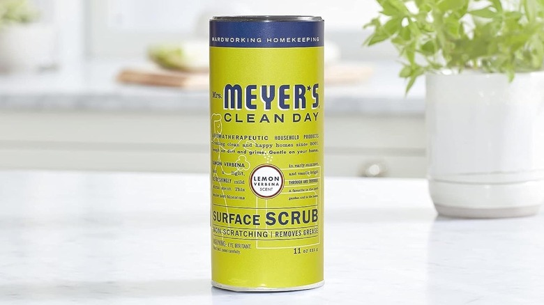 A jar of Mrs. Meyer's Clean Day Multi-Surface Scrub sits on a white countertop next to a potted plant