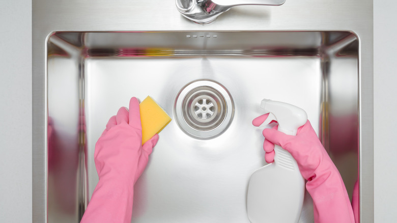 Woman in rubber gloves with sponge and cleanser cleaning a kitchen sink